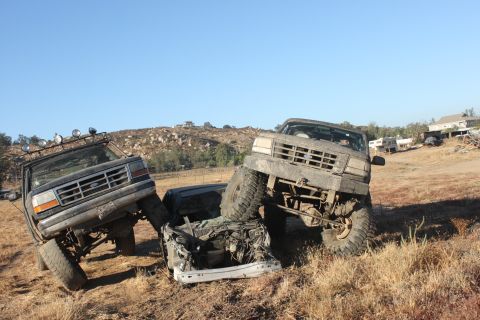 Two lifted trucks are on top of a wrecked car in a dry, grassy field