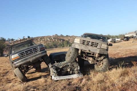 1992 Ford Bronco on the left, another Ford Bronco on the right, crushing a white car in a dry, grassy field