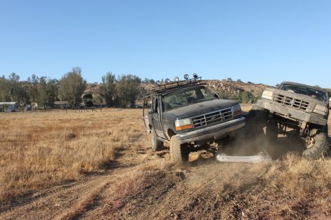 Two trucks are off-roading, one is stuck on a rock. The front truck has a roof rack with lights