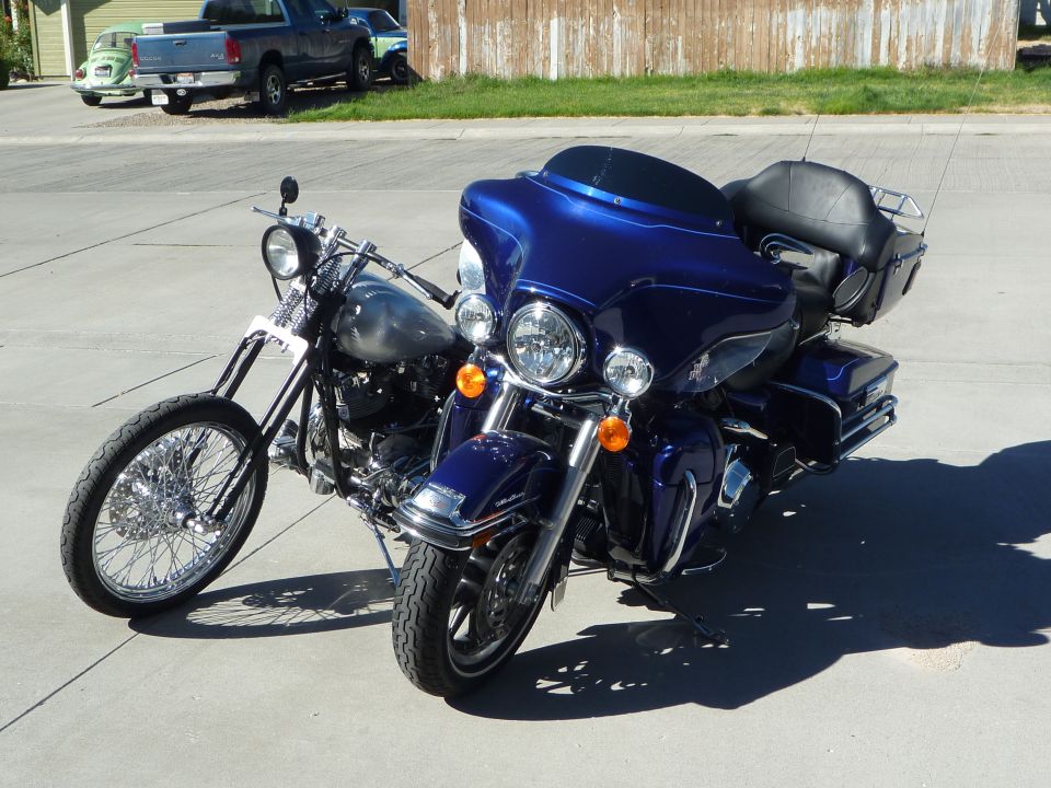 Two motorcycles parked on pavement. A blue touring motorcycle is in the foreground, with a chrome chopper behind it