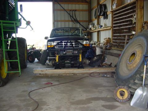 1992 Ford Bronco - Front view of a dark Ford Bronco raised on four jack stands inside a metal pole barn, with both front wheels removed and the yellow coil sp…