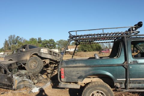 Two 1992 Ford Broncos, one green with a roof rack, the other brown and muddy, are piled up in a junkyard