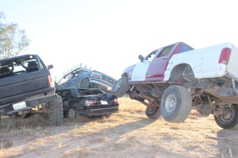 Several lifted trucks and a crushed car are in a dirt lot. One truck is white and maroon, another is black, an