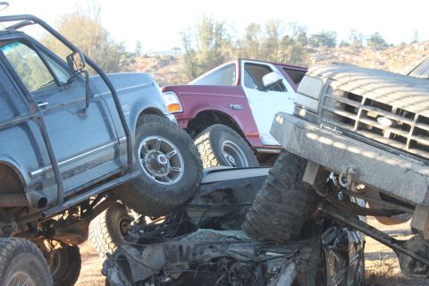 Several modified trucks are stacked on top of each other in a junkyard setting. One truck has a maroon cab and