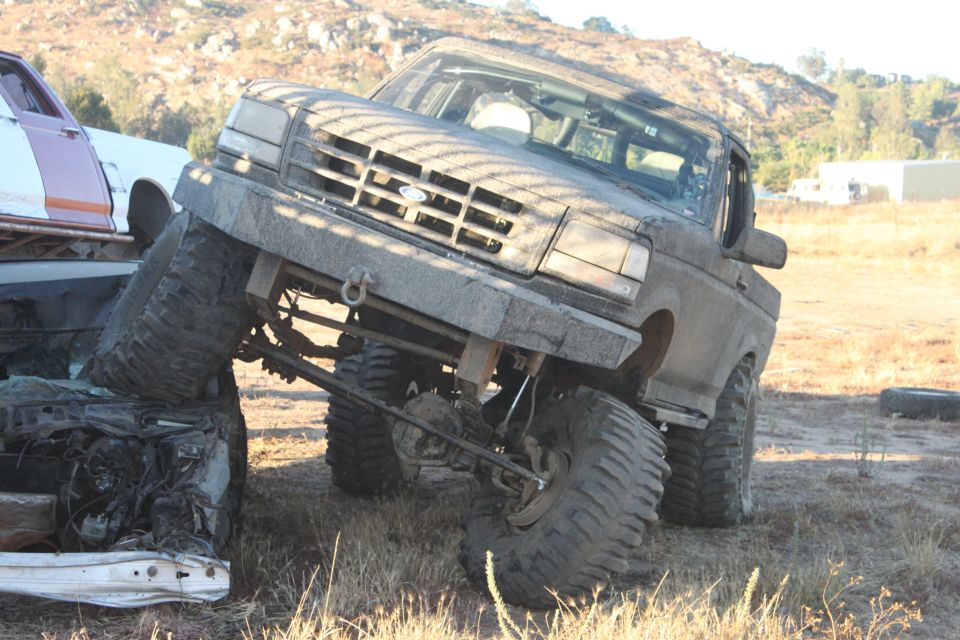 Mud-covered 1992 Ford Bronco with oversized tires, tilted on its side, crushing a car