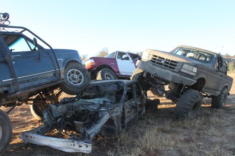 Three modified off-road trucks and a crushed car piled up. One truck is a 1992 Ford Bronco