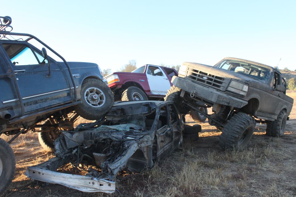 Three modified off-road trucks and a crushed car piled up. One truck is a 1992 Ford Bronco