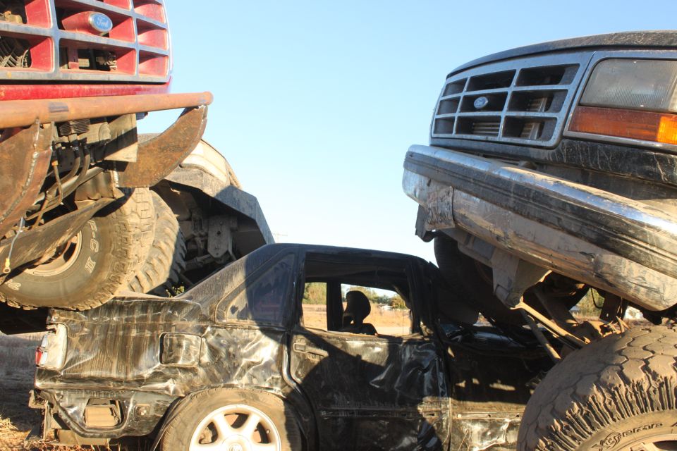 Crushed black sedan with a person inside, under a red truck and a dark SUV
