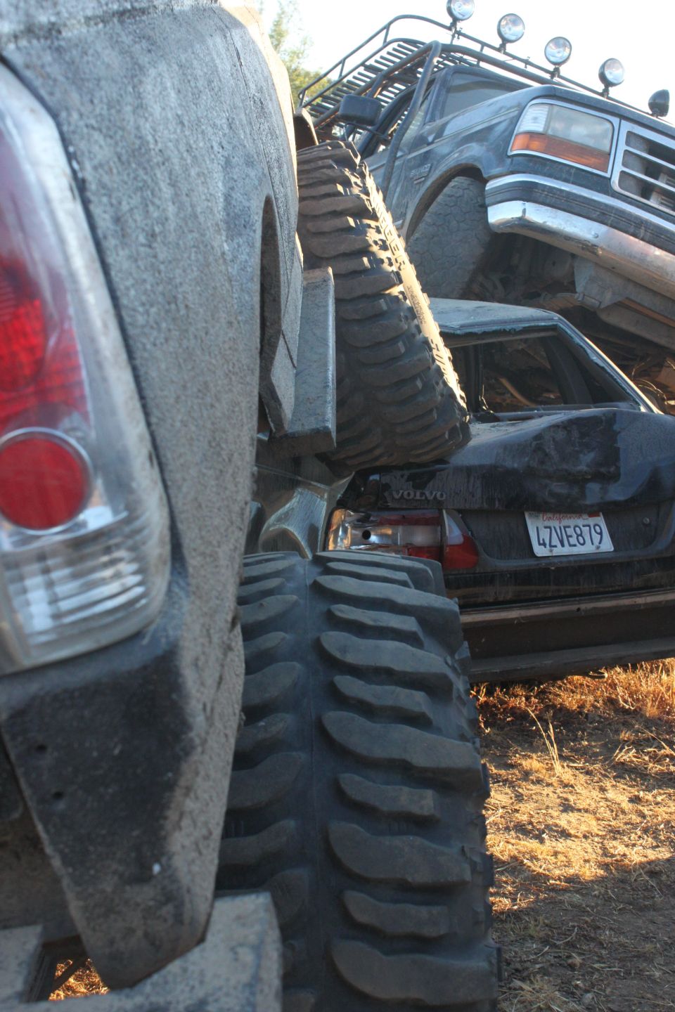 Truck with large tires in foreground, a 1992 Ford Bronco on top of a crushed Volvo