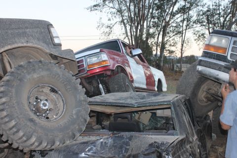 Red and white truck stuck on top of other vehicles, with a gray truck on the left and another truck on the rig