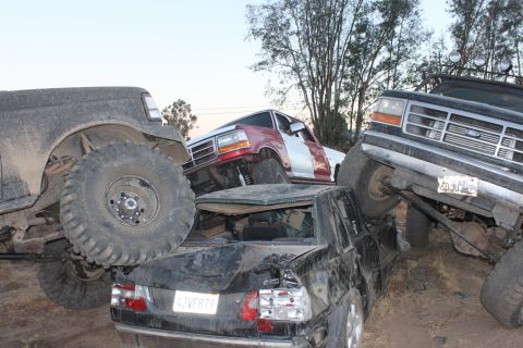 Three wrecked Ford trucks piled on top of each other, one with a red and silver two-tone paint job