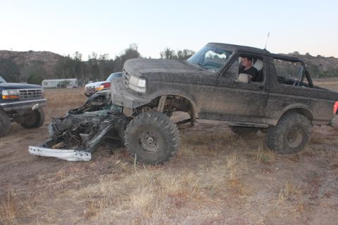 Muddy 1992 Ford Bronco with driver, next to a wrecked car. Another Ford truck is visible on the left