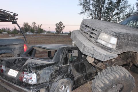 Big truck on top of a crushed car. Another truck is to the left