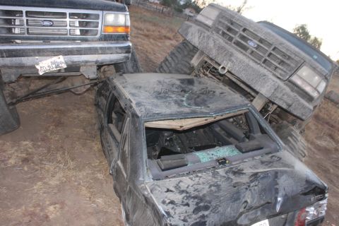 Two Ford trucks, one on top of a crushed sedan. The truck on the left appears to be a 1992 Ford Bronco. The tr