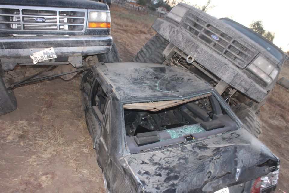 Two Ford trucks, one on top of a crushed sedan. The truck on the left appears to be a 1992 Ford Bronco. The truck on the right is also a Ford