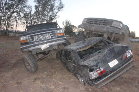 1992 Ford Bronco front end angled left, on top of a wrecked black sedan, with another vehicle behind