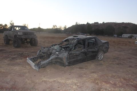 Heavily damaged black sedan in foreground, lifted pickup truck in background on dry grass field