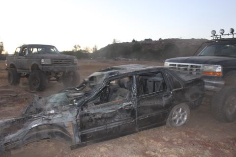 Wrecked black sedan in foreground, two lifted Ford trucks behind it