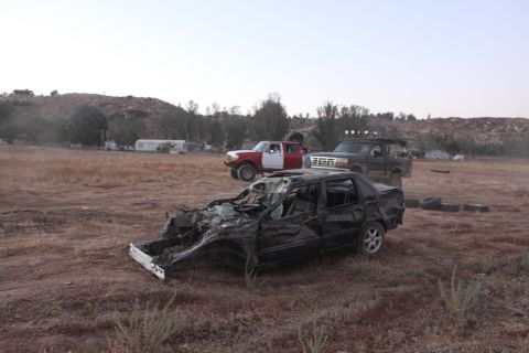 Wrecked black sedan in foreground, two trucks behind it on dry ground