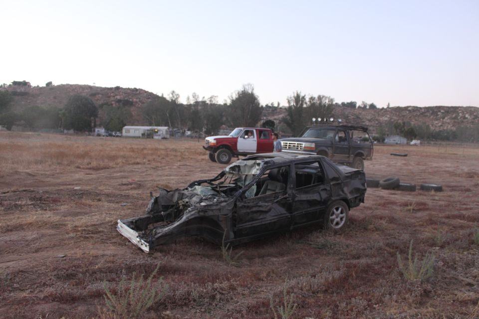 Wrecked black sedan in foreground, two trucks behind it on dry ground