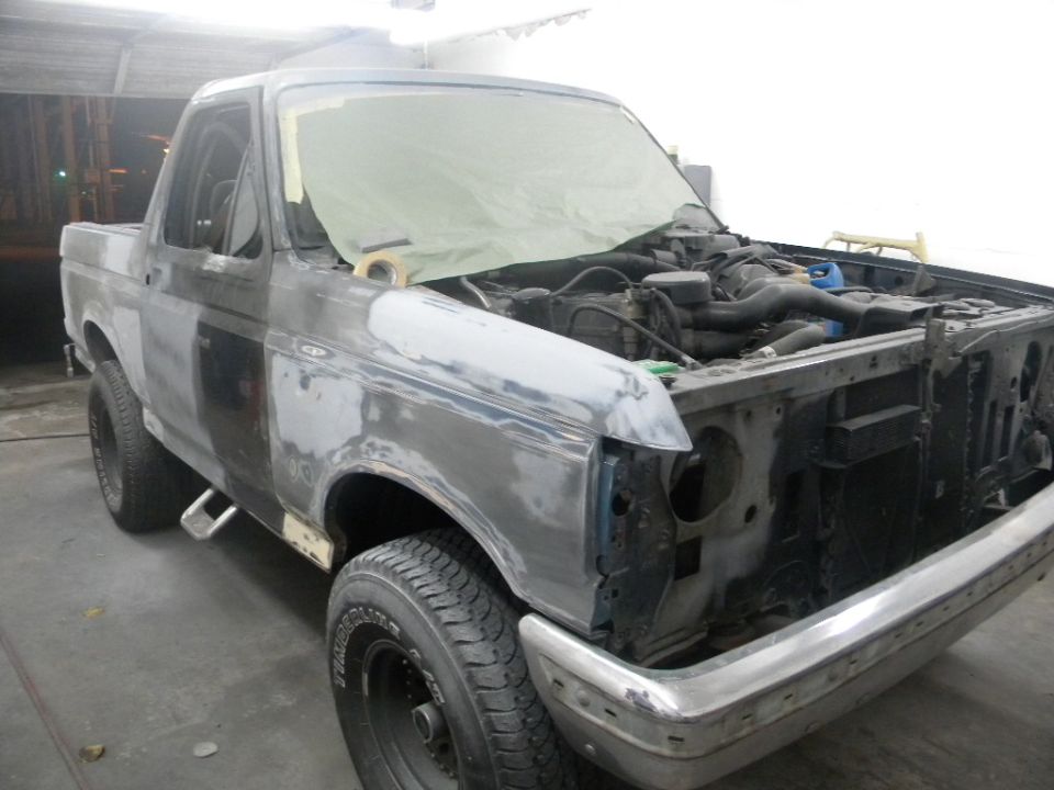 1987 Ford Bronco undergoing bodywork, hood open, engine exposed, primer finish