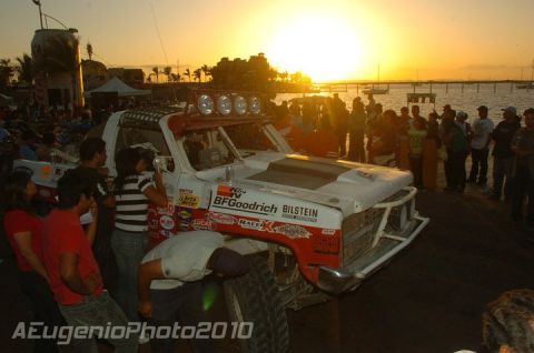 1990 Ford Bronco - white off-road race truck with roof lights and BFGoodrich decals surrounded by a crowd at a waterfront event during sunset.