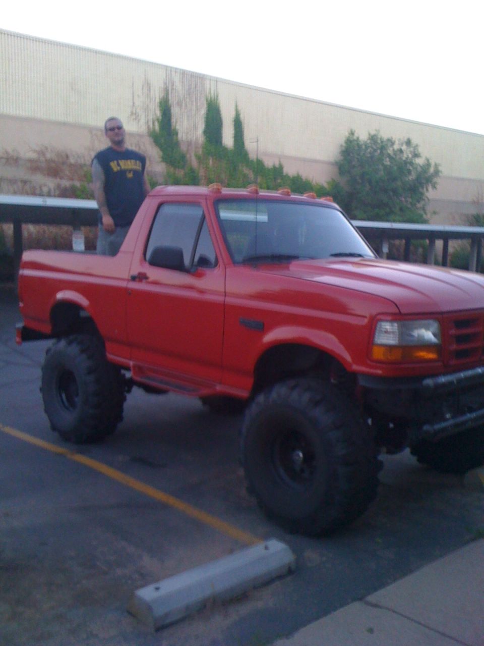 Red lifted Ford truck on large mud tires with man standing beside it in parking lot