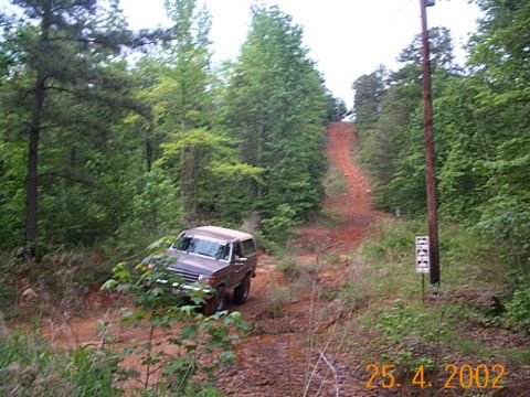 1986 Ford Bronco - A brown 1986 Ford Bronco navigates a muddy, uphill dirt road surrounded by trees.
