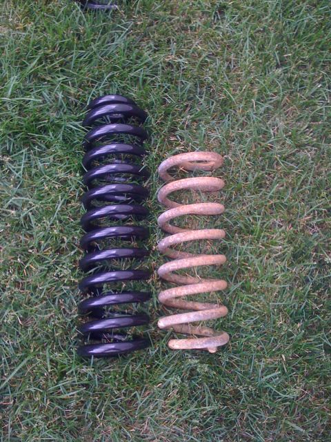 1992 Ford Bronco - Comparison of two coil springs, one new and black, the other old and rusted, resting on a grassy surface.
