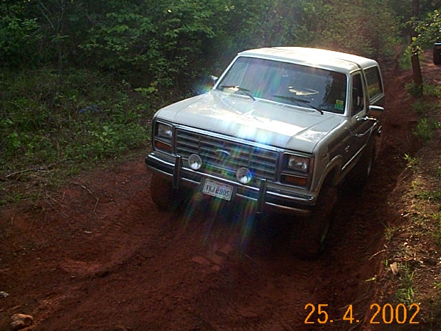 Two-tone silver Ford Bronco with a brush guard on a red-dirt woodland trail, front three-quarter view