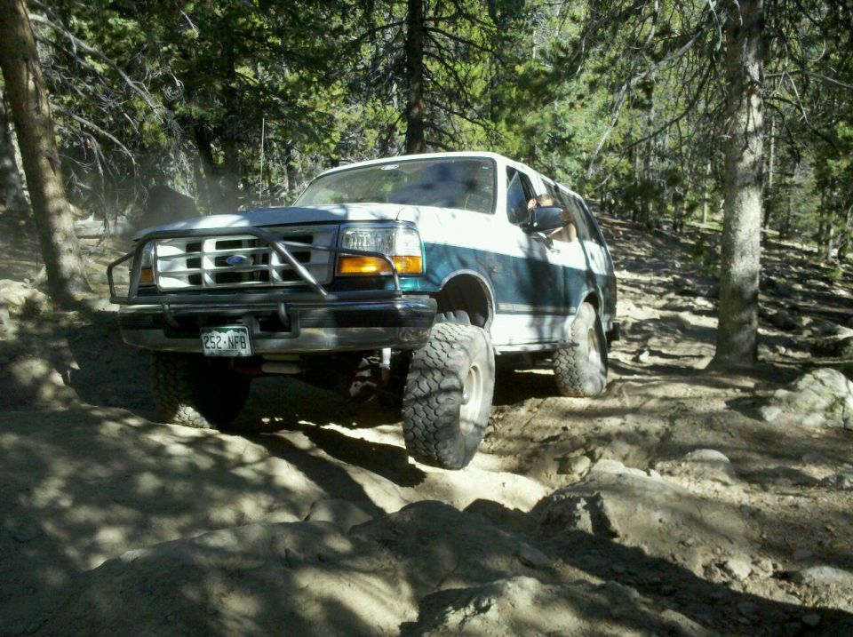 1996 Ford Bronco with aftermarket bumper and large tires driving on a rocky, wooded trail