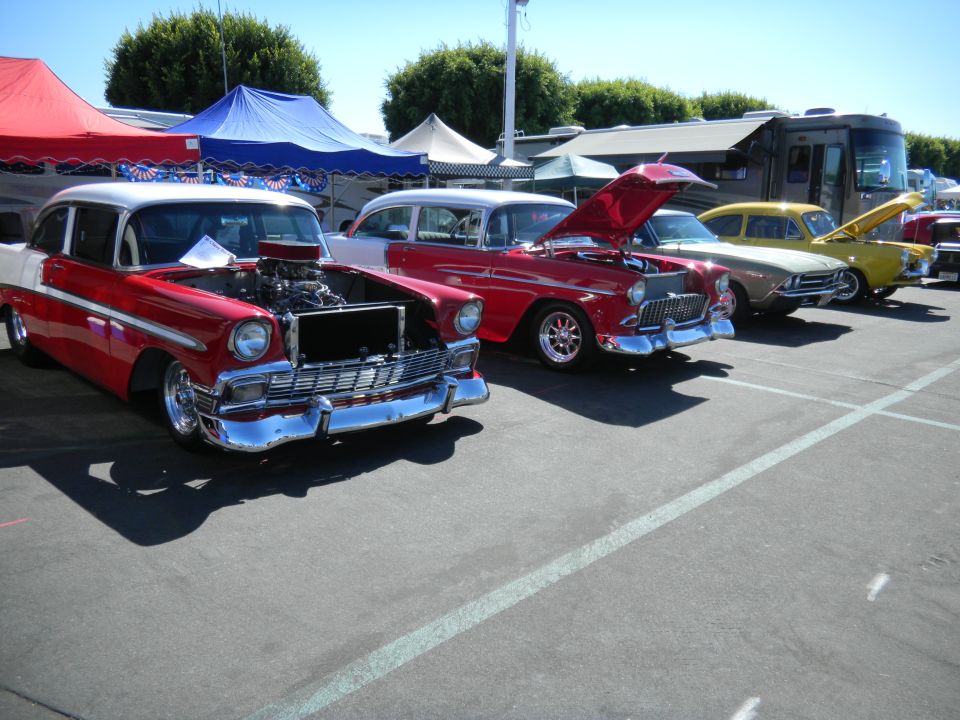 Several classic cars parked in a lot, some with hoods open. Tents and an RV are in the background