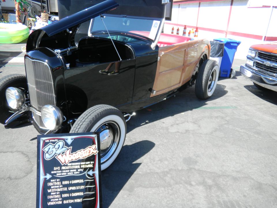 Black hot rod with wood paneling and red interior, hood open, parked next to a blue trash bin and an orange truck