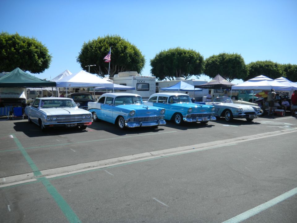 Several classic cars parked in a lot under tents, with trees and a clear blue sky in the background