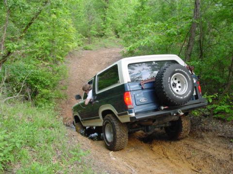1986 Ford Bronco - A Ford Bronco navigates a muddy off-road trail surrounded by dense green foliage.