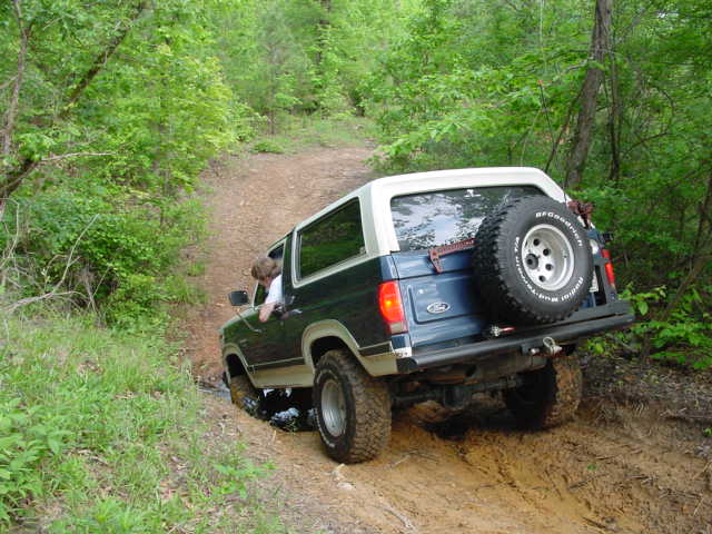 Green Ford Bronco with white top and tailgate spare descending a muddy woods trail