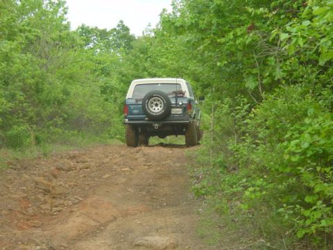 1986 Ford Bronco - A blue Ford Bronco drives on a dirt road surrounded by lush green trees.