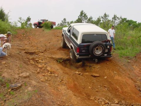 1986 Ford Bronco - A Ford Bronco navigates a steep, muddy off-road trail with people and a dog watching.