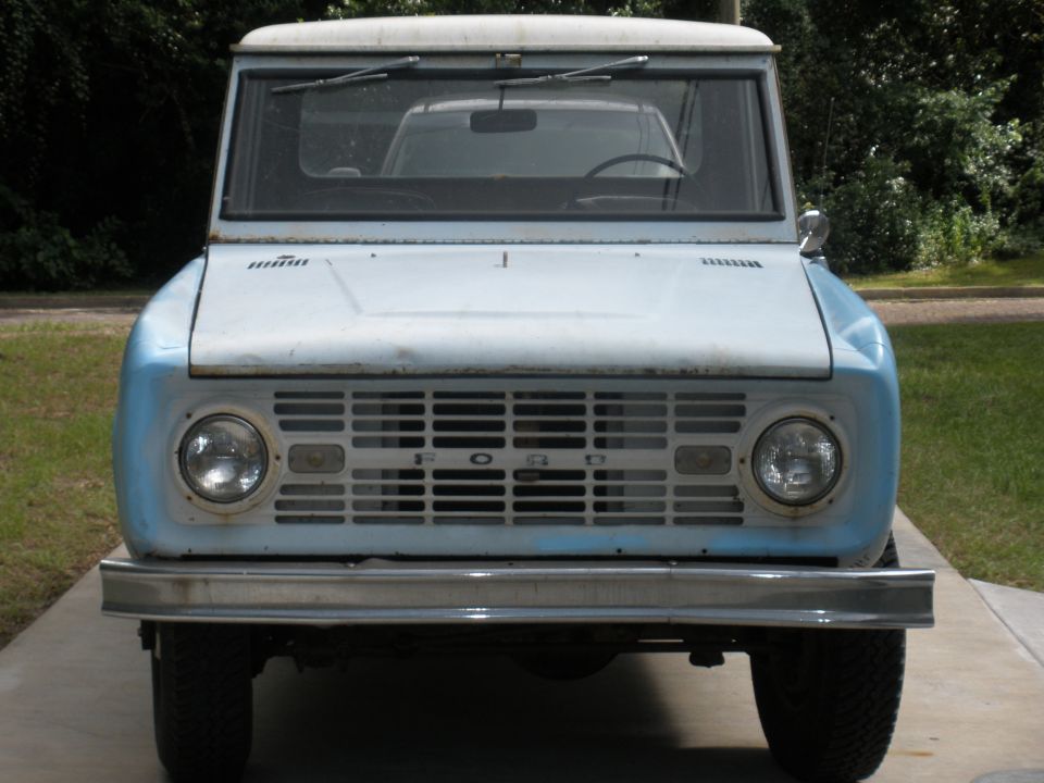 Front view of a light blue first-generation Ford Bronco with a white roof. The grille has 'FORD' spelled out