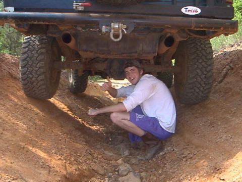 1986 Ford Bronco - A person poses under the rear of a lifted SUV, giving a thumbs up while off-roading.
