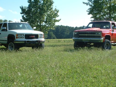 1994 GMC Suburban - A white 1994 GMC Suburban and a red pickup truck are parked in a grassy field under a clear blue sky.