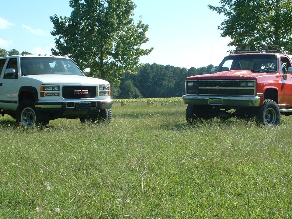 Lifted white GMC truck and red Chevy K5 Blazer facing camera in a grassy field
