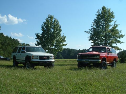 1994 GMC Suburban - Two SUVs, a white 1994 GMC Suburban and a red Chevrolet K5 Blazer, sit in a grassy field under a blue sky.