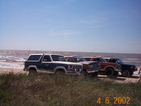 1986 Ford Bronco - Several SUVs are parked on a beach with the ocean and sky visible.
