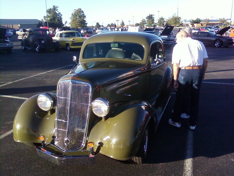 Olive green vintage coupe with chrome grille and round headlights, man standing next to it. Other cars in background