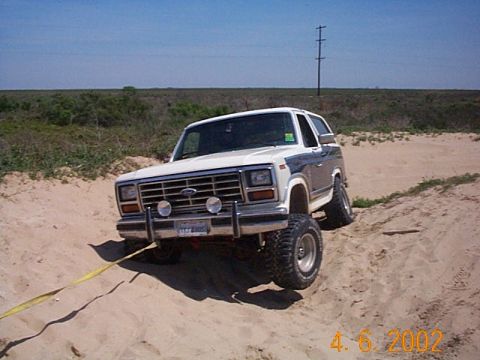 1986 Ford Bronco - Ford Bronco with off-road tires is stuck in the sand, attached to a tow strap.