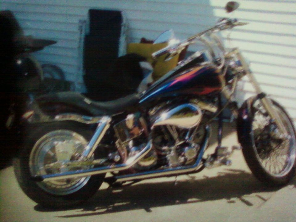 Custom cruiser motorcycle with ape-hanger bars and chrome exhaust parked by a white-sided building