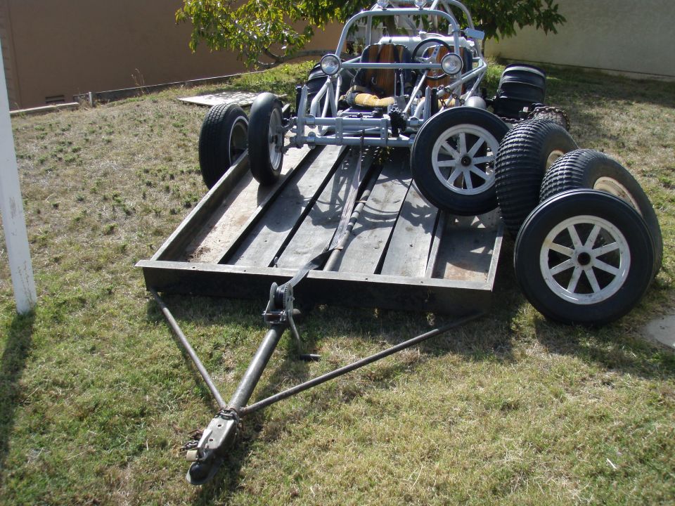 Tube-frame sand rail buggy loaded on a wood-deck utility trailer with spare wheels on the grass