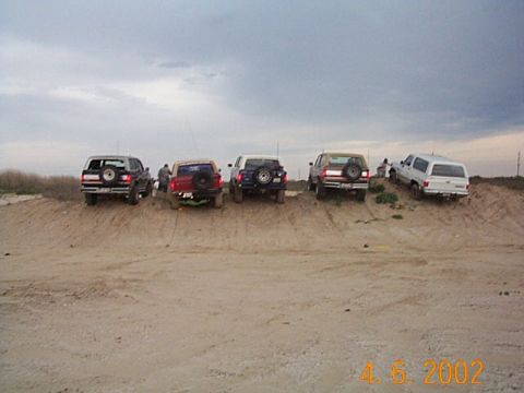 1986 Ford Bronco - Multiple SUVs are positioned on a sandy terrain under an overcast sky.