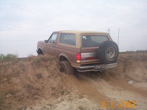 1986 Ford Bronco - This 1986 Ford Bronco is seen from the rear, navigating a dirt path with its spare tire mounted externally.